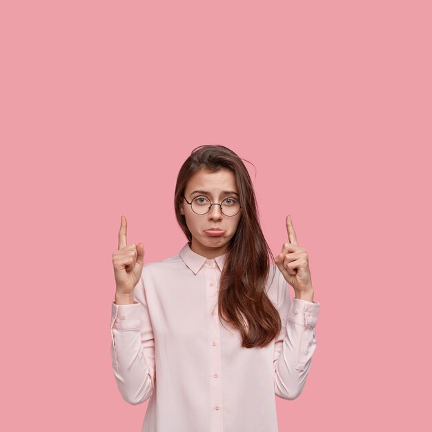 Vertical shot of unhappy young woman purses lower lip, indicates with both index fingers