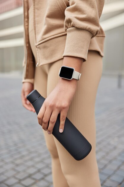 Vertical shot of unrecognizable woman dressed in beige sportswear wears smartwatch holds bottle of fresh water takes break after workout. Faceless sporty female model feels thirsty during training