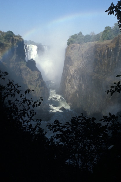 Vertical shot of a waterfall flowing down from tall hills under a blue sky with a rainbow