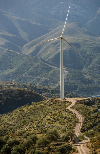 Vertical shot of a white wind fan standing on a green field behind the mountains