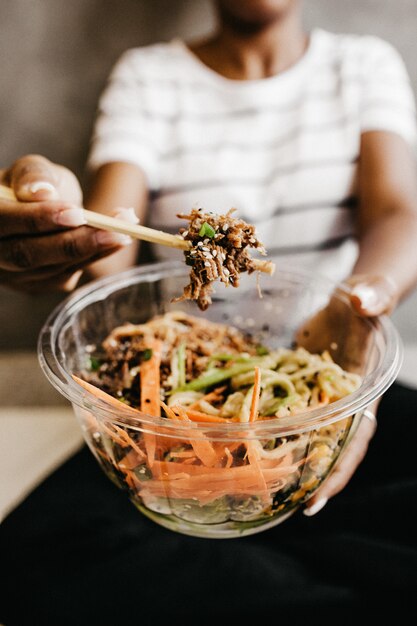 Vertical shot of a woman holding a clear plastic bowl with vegetable salad and chopsticks