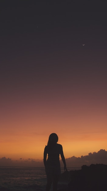 Vertical shot of a woman in silhouette standing on a cliff near the sea during sunset