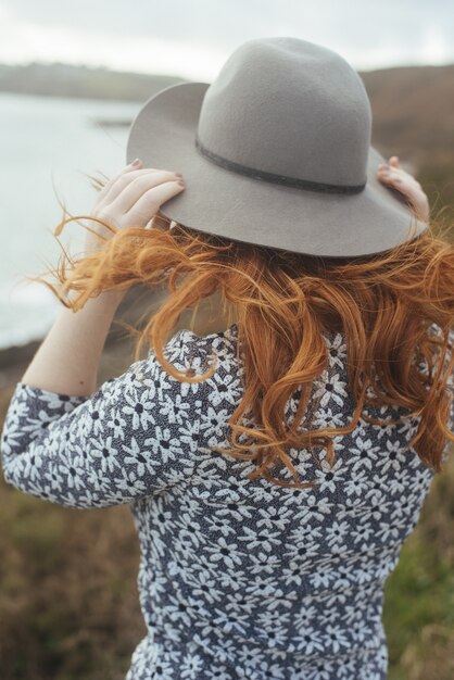 Vertical shot of a woman wearing a hat with the sea and trees on the distance