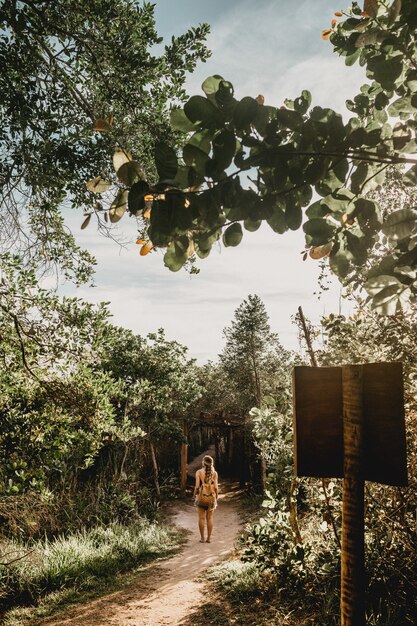 Vertical shot of a woman with a backpack walking on a forest path barefoot