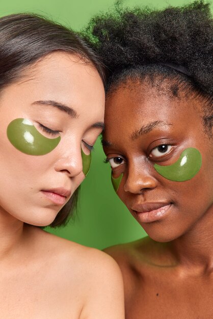 Vertical shot of women with different skin stand bare shoulders against vivid green wall apply hydrogel patches under eyes reduce fine lines undergo beauty treatments. Skin care