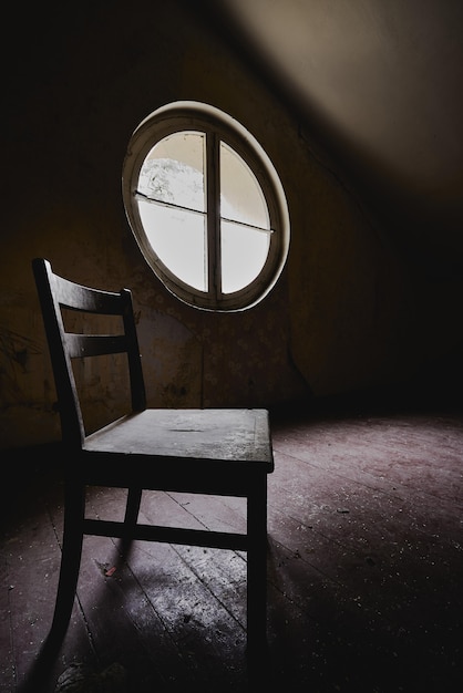 Vertical shot of a wooden chair in a dark room with a round window - concept of isolation