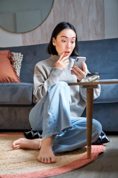 Vertical shot of young asian girl looks surprised at her mobile phone reads amazing news notificatio