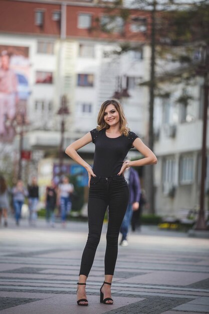 Vertical shot of a young blonde female in black pants, blouse and high heels posing in the street