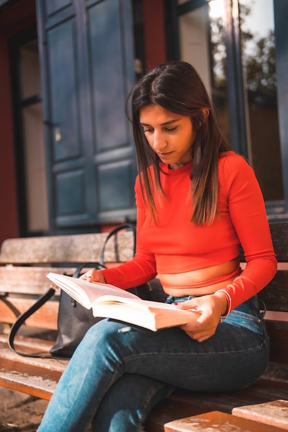Vertical shot of a young Caucasian brunette in a red stylish blouse reading a book at a bench park
