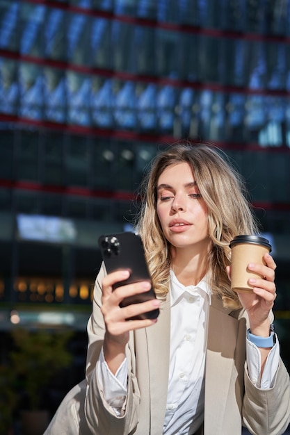 Vertical shot of young corporate woman drinks her coffee and looks at mobile phone texts message sta