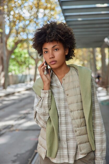 Vertical shot of young curly haired woman concentrated into distance talks on mobile phone enjoys phone conversation