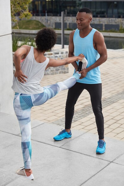 Вертикальный снимок молодой темнокожей женщины с болит в спине, поднимает ноги, вместе с тренером делает упражнения на растяжку, позирует на улице. Единение, спорт, концепция обучения. Черный парень помогает с тренировкой