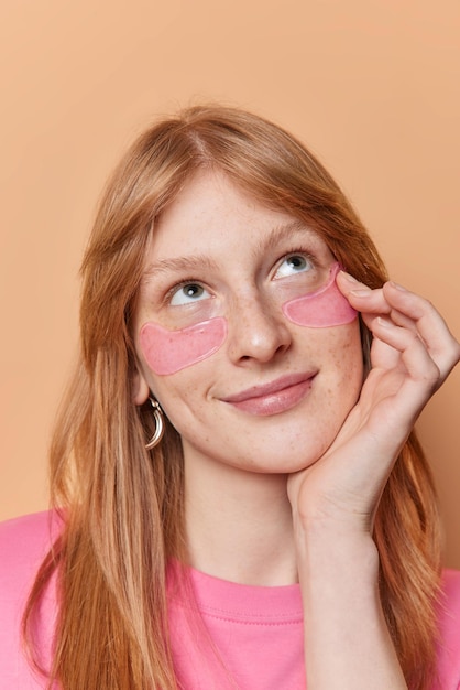 Vertical shot of young European woman applies hydrogel patches under eyes focused above has long red hair thinks about something isolated over beige background Beauty procedures and skin care