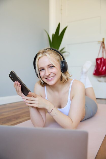 Free photo vertical shot of young fitness woman in headphones lying on rubber mat relaxing after workout