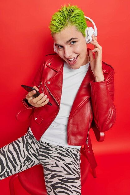 Vertical shot of young good looking woman with trendy hairstyle listens music via stereo headphones holds mobile phone sits on comfortable chair isolated over red background. Technology concept