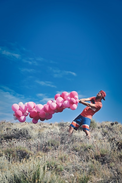 Вертикальный снимок молодого мужчины, несущего кучу розовых шаров на поверхности голубого неба