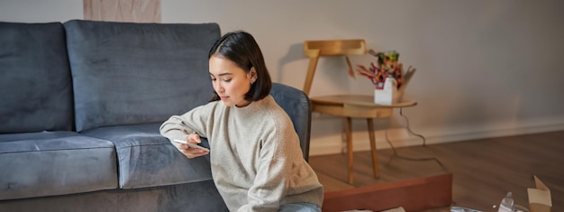 Vertical shot of young woman in cozy home working on laptop using smartphone and drinking coffee