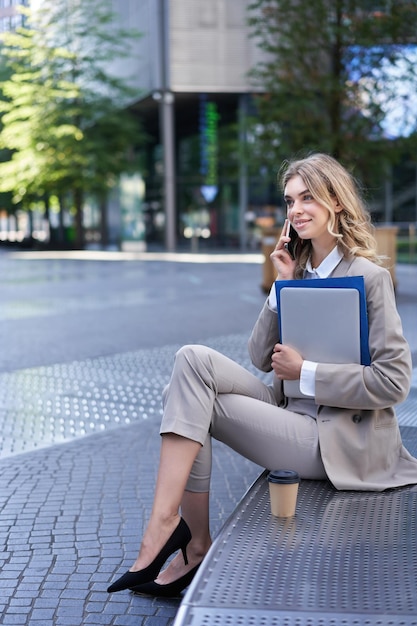 Ripresa verticale di una giovane donna seduta vicino a un edificio per uffici che tiene laptop e cartelle con docum di lavoro