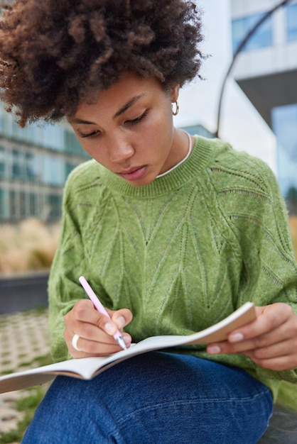 Free photo vertical shot of young woman with curly hair writes organisation plan in textbook for education uses pen processes information for university course work wears sweater and jeans poses outdoors