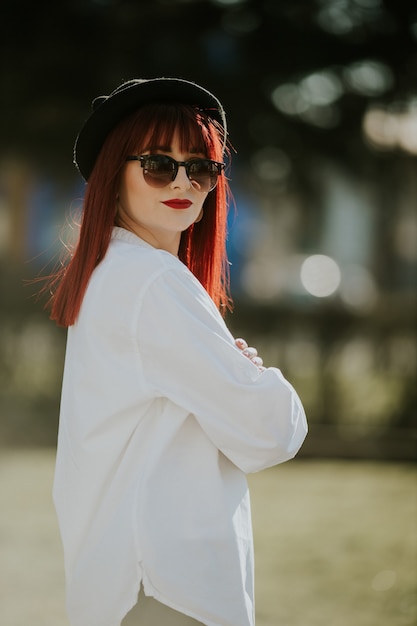 Vertical side view of a young smiling female with red hair, sunglasses, and a hat posing in a park