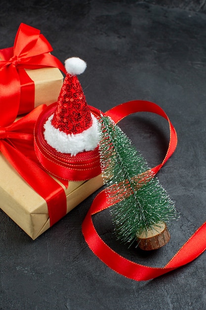 Вертикальный вид красивых подарков с красной лентой и новогодней елкой в шапке санта-клауса на темном столе