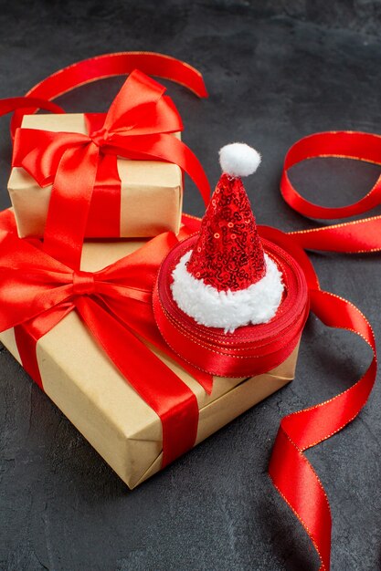 Вертикальный вид красивых подарков с красной лентой и шляпой санта-клауса на темном фоне
