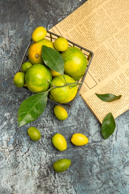 Vertical view of black basket with fresh green tangerines and kumquats on newspapers on gray background