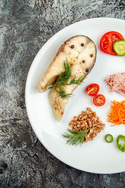 Vertical view of boiled fish buckwheat served with vegetables green on a white plate on ice surface