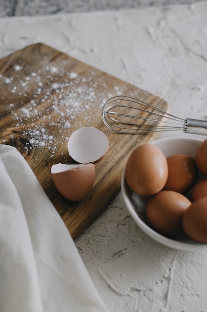 Vertical view of a bowl of eggs and a silver whisk next to a chopping board on a white surface