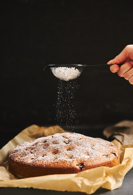 Vertical view of a Cherry Cake with sugar powder and ingredients on the side on a black background