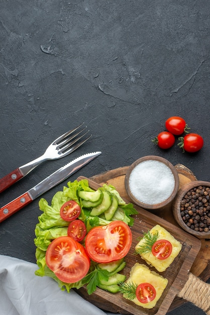 Free photo vertical view of chopped and whole fresh vegetables cheese on cutting board and spices cutlery set on black surface