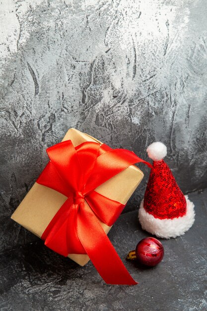 Vertical view of christmas gift with red ribbon standing on gray wall and santa claus hat and decoration accessory on dark background