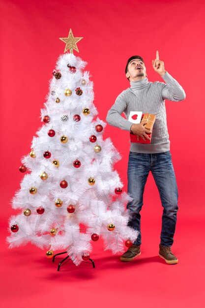 Vertical view of confused handsome man standing near the decorated white xmas tree