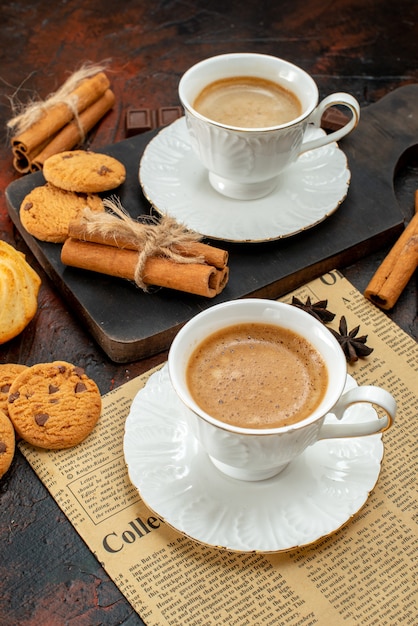 Vertical view of cups of coffee on wooden cutting board and an old newspaper cookies cinnamon limes chocolate bars on dark background