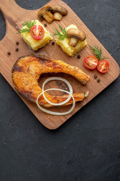 Vertical view of delicious fried fish meal and mushrooms tomatoes greens on wooden cutting board on black surface