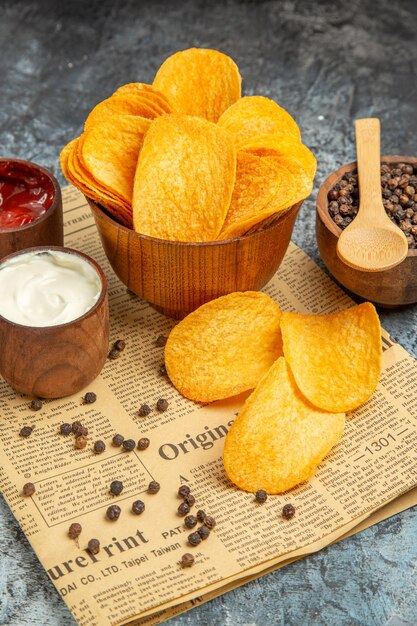 Vertical view of delicious homemade chips and pepper bowl mayonnaise ketchup and sauce with spoon on newspaper on gray background