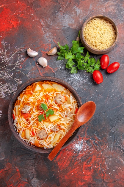 Vertical view of delicious noodle soup with chicken and uncooked pasta in a small brown bowl and spoon garlic tomatoes and greens on the dark background