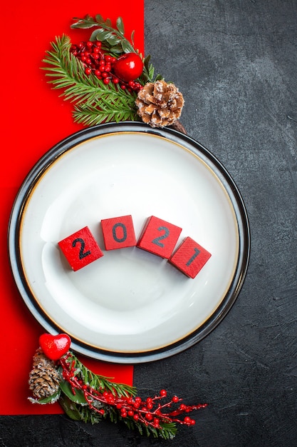 Vertical view of dinner plates with numbers and fir branches with decoration accessory conifer cone on a red napkin on a dark background