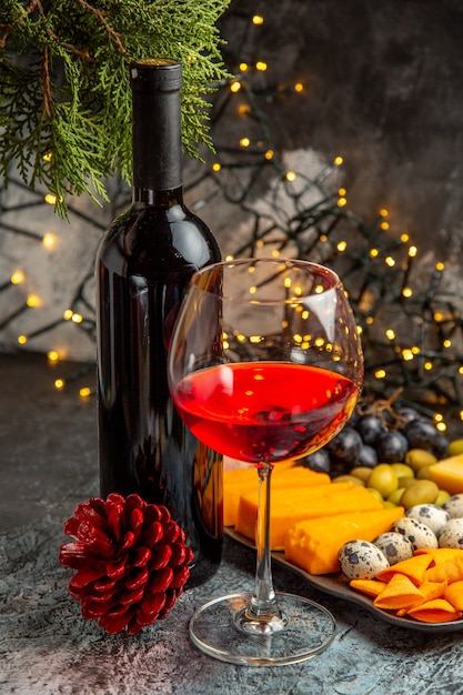 Free photo vertical view of dry red wine in a glass and in a bottle next to snack and conifer cone on gray background