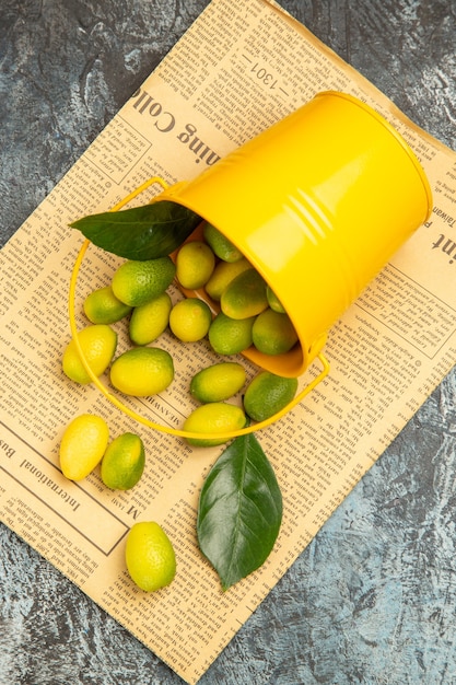 Free photo vertical view of fallen yellow bucket with fresh kumquats on newspapers on gray background