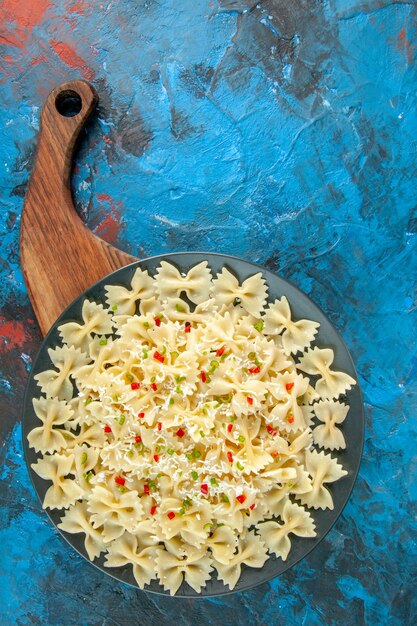 Vertical view of farfalle pastas with vegetables on a black plate on wooden cutting board on blue background