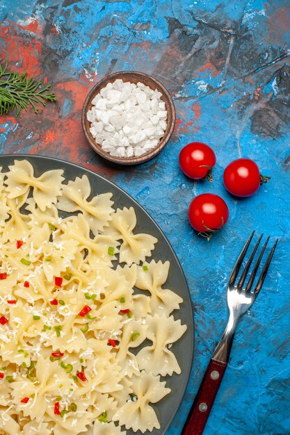 Vertical view of farfalle pastas with vegetables tomatoes salt and fork fir branches on blue background