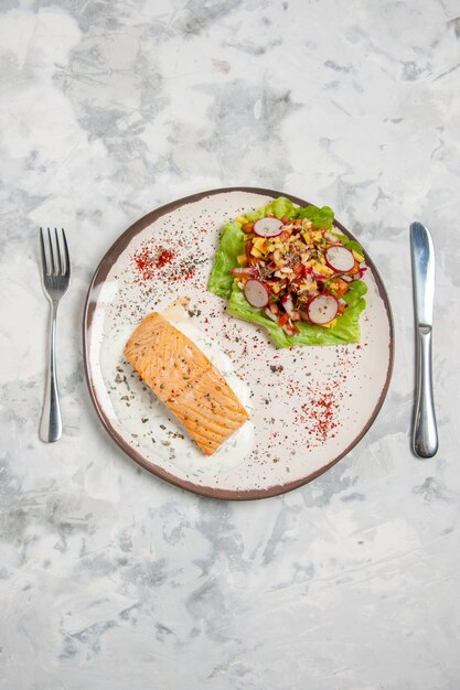 Vertical view of fish meal and delicious salad on a plate and cutlery set on stained white surface