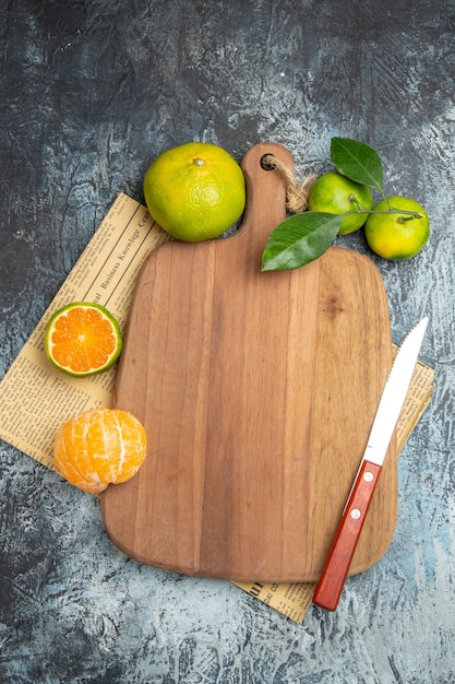 Free photo vertical view of fresh citrus fruits with leaves around the wooden cutting board cut in half forms and knife on newspaper on gray background