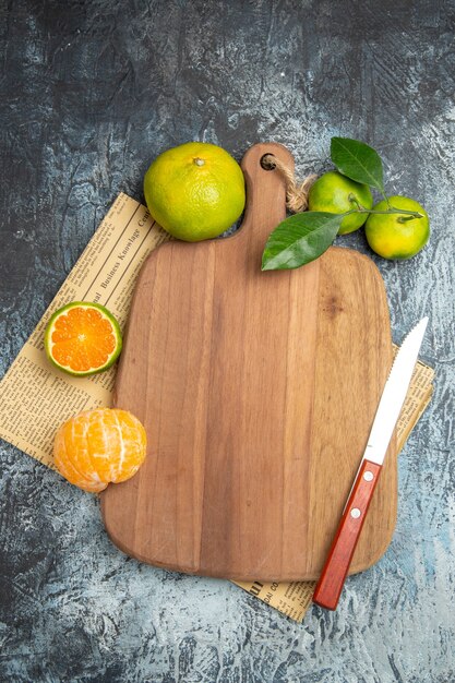 Vertical view of fresh citrus fruits with leaves around the wooden cutting board cut in half forms and knife on newspaper on gray background