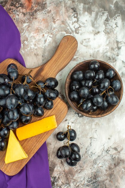 Vertical view of fresh delicious black grape bunch and cheese on wooden cutting board and in a brown pot on purple towel on mixed color background