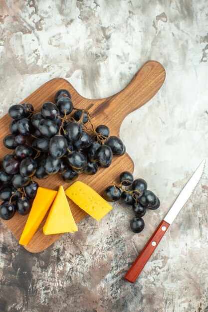 나무 커팅 보드에 있는 신선한 맛있는 검은 포도 다발과 다양한 종류의 치즈와 혼합 색상 배경에 있는 칼의 수직 보기
