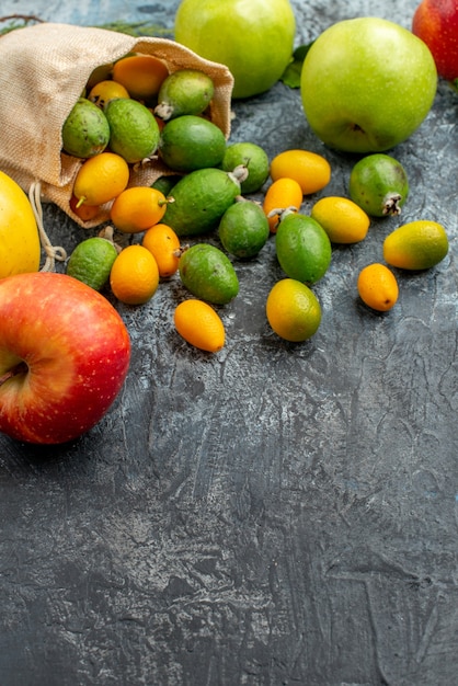 Foto gratuita vista verticale di kumquat freschi all'interno e all'esterno di una piccola borsa bianca caduta e mele verdi gialle rosse sul tavolo grigio