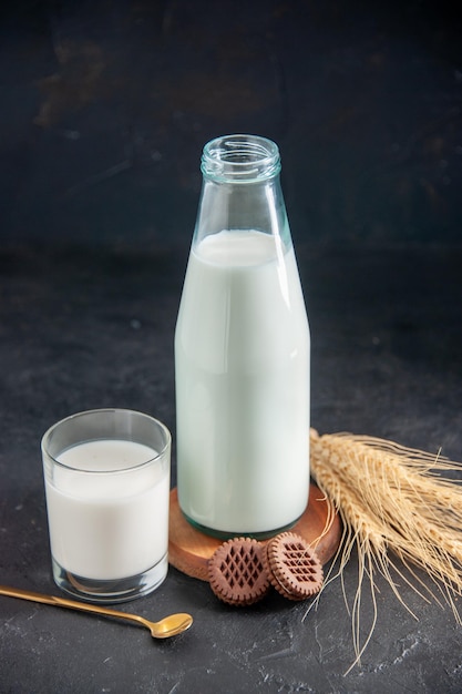 Vertical view of a fresh milk in a bottle and glass cookies and golden spoon on small brown wooden tray spikes on black wave background