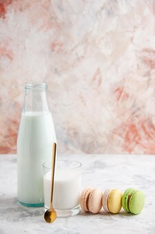 Vista verticale del latte fresco in una tazza di vetro e bottiglia aperta accanto a deliziosi macarons colorati su sfondo colori pastello